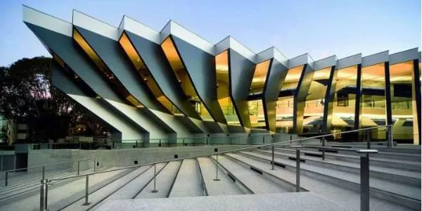 Modern building with angular metal fins and illuminated glass panels above concrete steps with metal handrails at dusk
