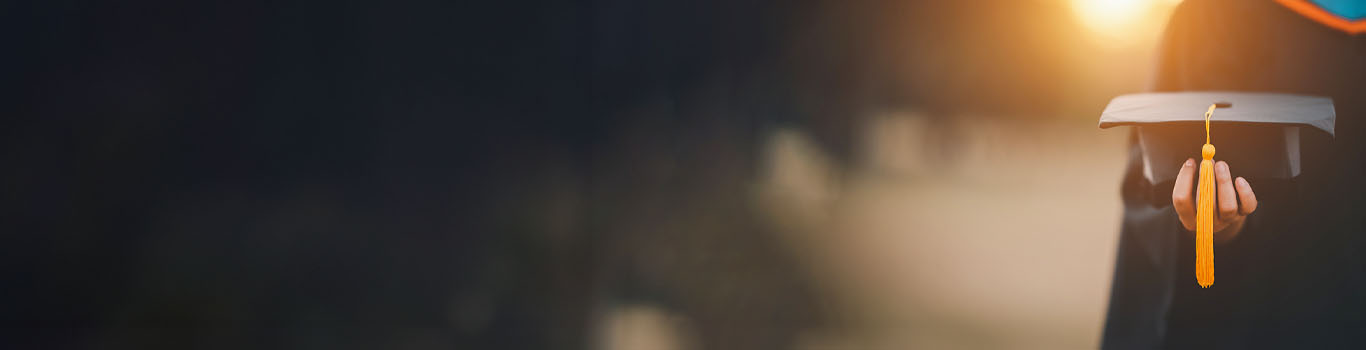 Graduate holding grad cap with a yellow tassel hanging down with the sun behind them
