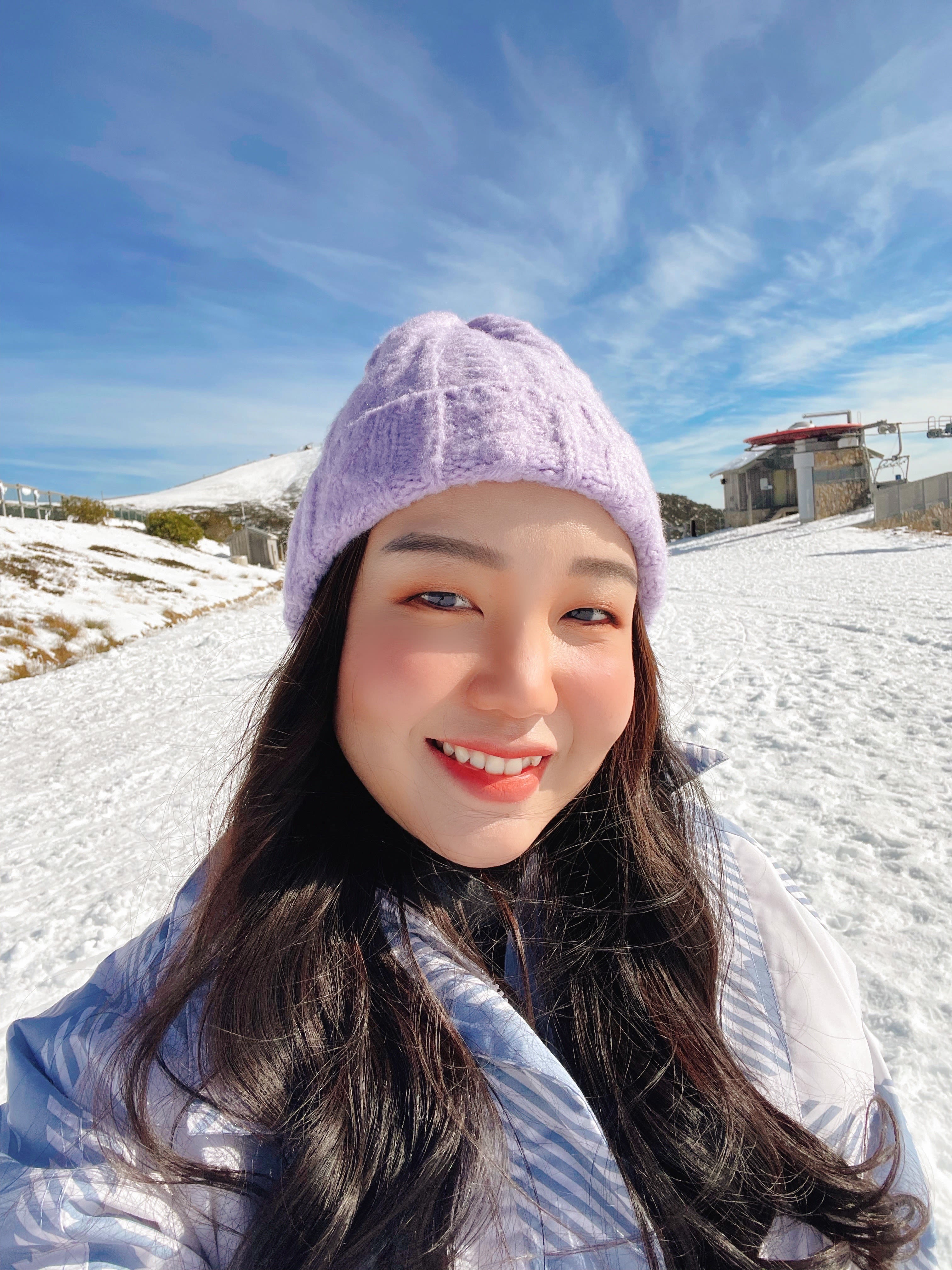 A person wearing a purple knit hat and a winter coat smiles while standing in a snowy landscape. The sky is blue with wispy clouds, and there are buildings and a snow-covered hill in the background.