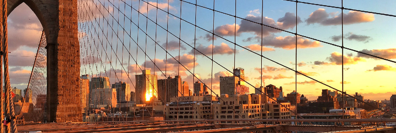 View of a city skyline at sunset from the Brooklyn Bridge, featuring the bridges cables in the foreground and buildings silhouetted against a colorful sky with clouds.