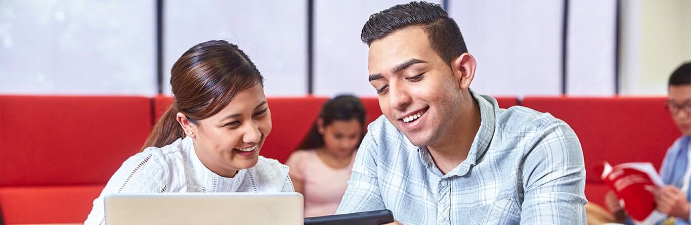 Two people are sitting together, smiling and looking at a tablet. The woman on the left has dark hair tied back, and the man on the right has short dark hair. In the background, another person is reading a book. They are seated indoors.