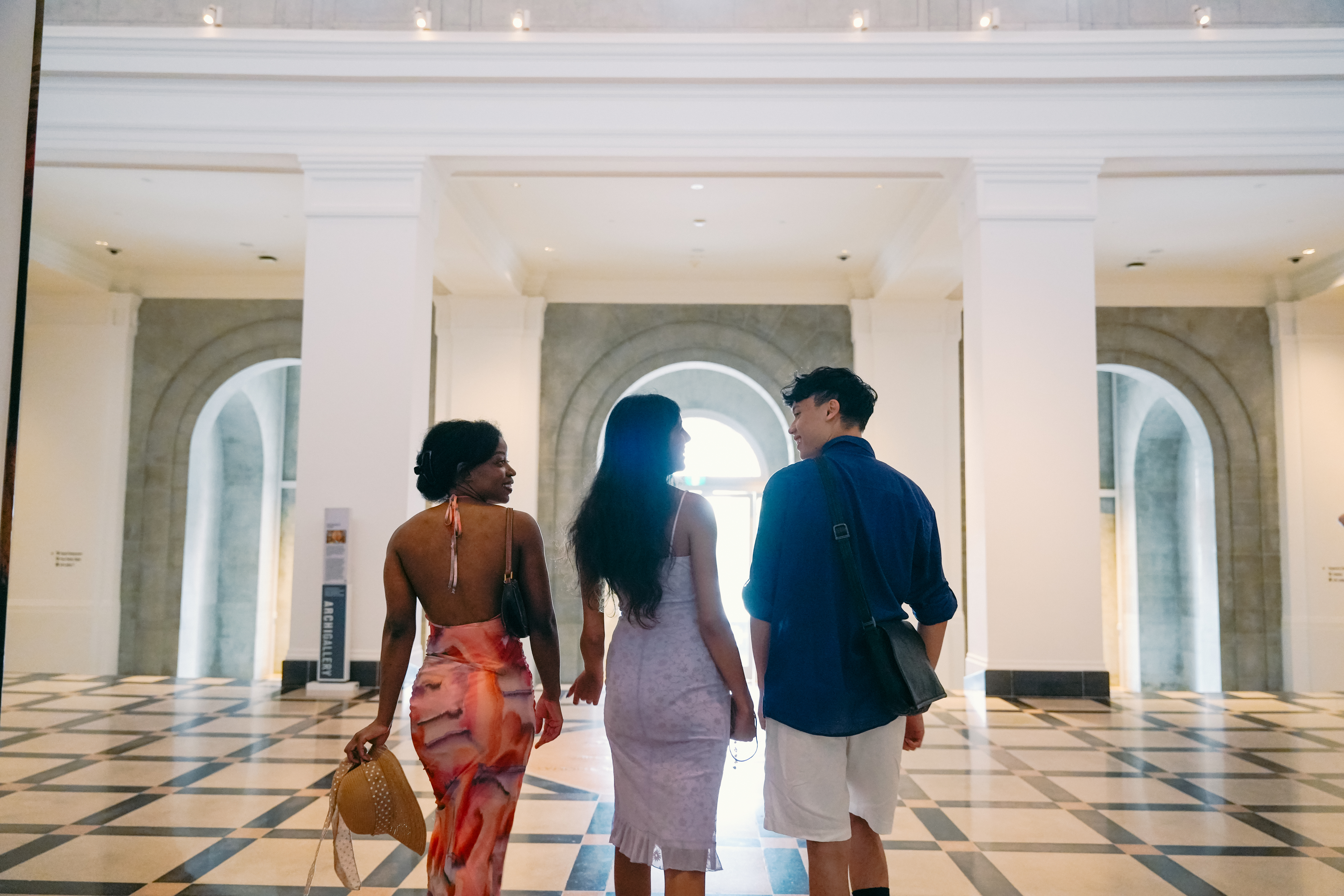 Three people walking through an elegant white hallway with arched windows and a geometric patterned floor, backlit by natural light