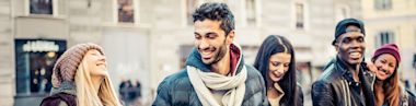 A diverse group of young adults stands outdoors in a city, smiling and laughing together. They wear casual winter clothing, including jackets, scarves, and hats, and appear to be enjoying each others company.