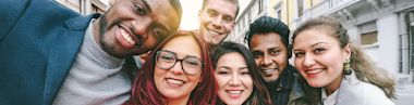 A group of six young adults of diverse backgrounds smiling and posing closely together for a selfie outdoors on a city street.