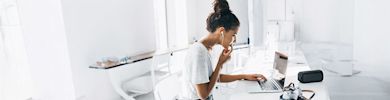 A person wearing earphones is seated at a desk working on a laptop in a bright, modern office space. There are several gadgets on the desk, and the background features sleek, white decor.