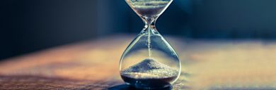 An hourglass sits on a wooden surface with sand flowing from the top chamber to the bottom, illuminated by soft light from the side. The background is dark and out of focus.