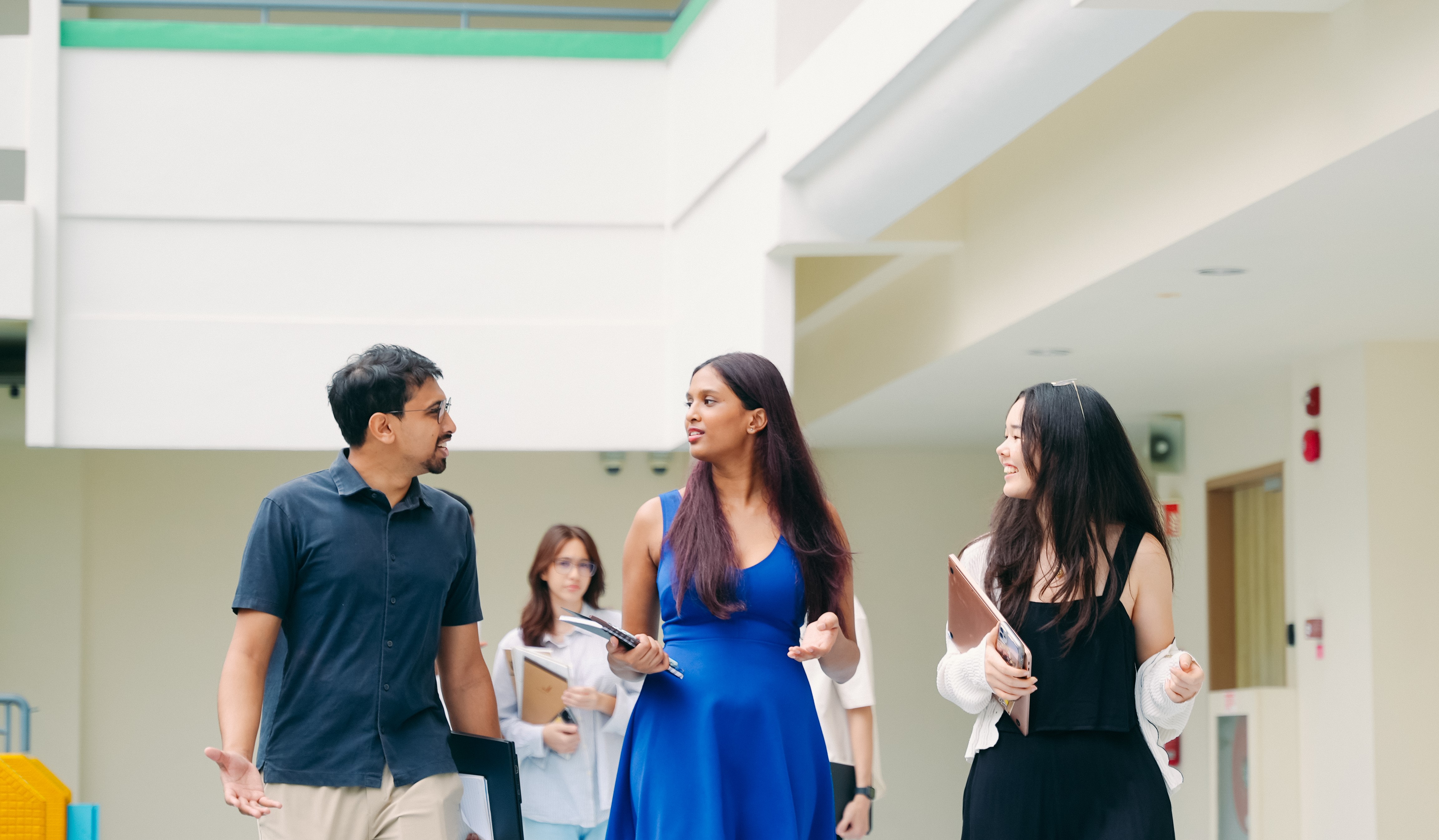 Students walking and conversing in a bright hallway, carrying notebooks and papers, wearing casual and business attire