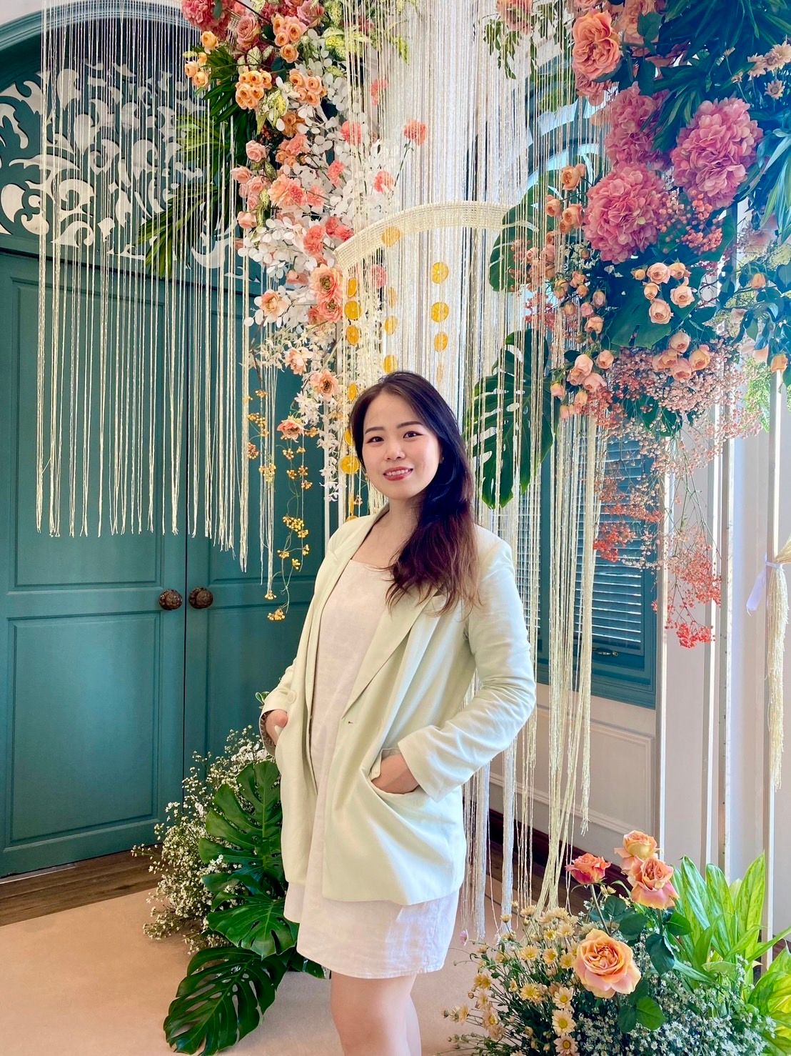 A person stands smiling indoors, wearing a light green blazer over a white dress. Behind them is a decorative floral arrangement with pink, orange, and white flowers, and green leaves, set against blue double doors.