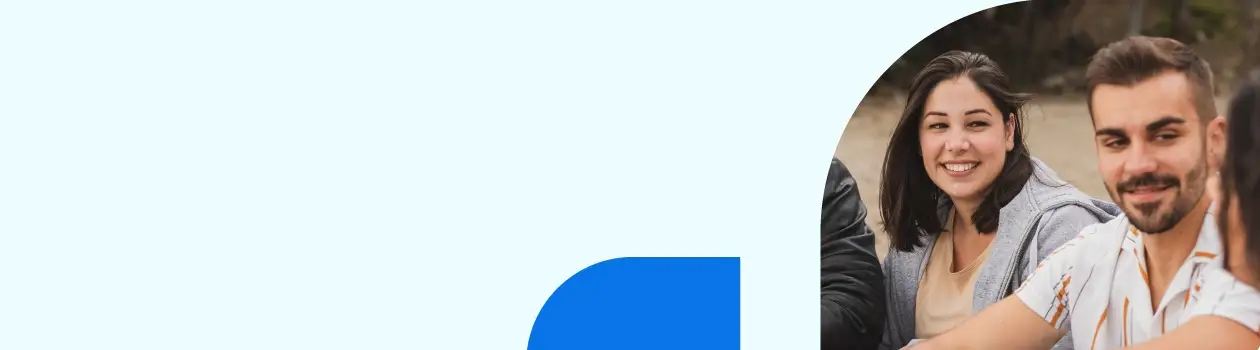 A group of people sitting together outdoors, smiling and talking. The background is blurred, and the left side of the image has a light blue area with a blue geometric shape.