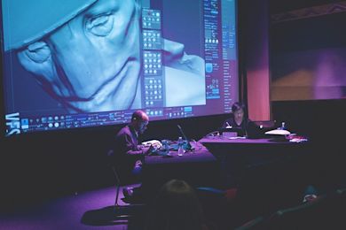 Two people work on laptops in a dimly lit room. A digital art projection of a humanoid face with blue tones is displayed on a large screen behind them, showcasing a software interface with various tools and icons.