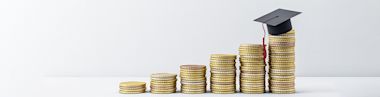 Stacks of coins arranged in ascending order, with the tallest stack topped by a graduation cap, symbolizing the increasing cost or value of education.