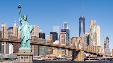 A cityscape featuring a river with a bridge, surrounded by tall skyscrapers under a blue sky with clouds. The buildings display a mix of architectural styles, with modern glass structures and historic brick designs.