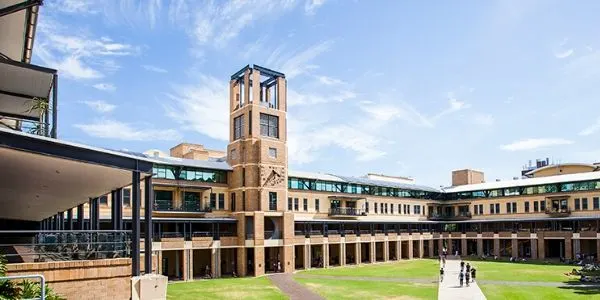 Modern university campus building with tan brick walls, glass corridors, and distinctive clock tower under blue sky