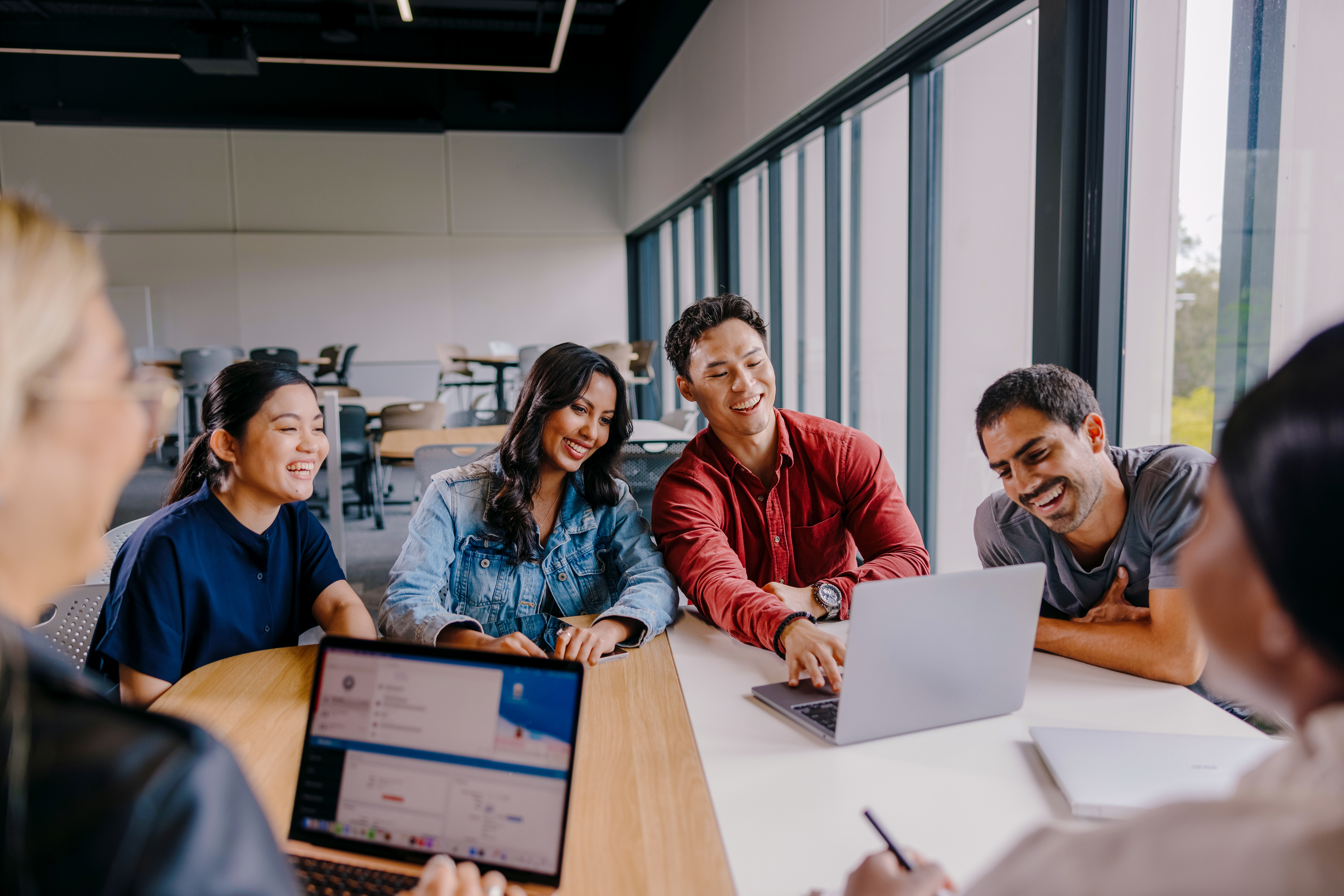  A diverse group of five young adults smiling and looking at a laptop screen around a light-colored conference table in a modern office space with large windows. All appear engaged and happy.