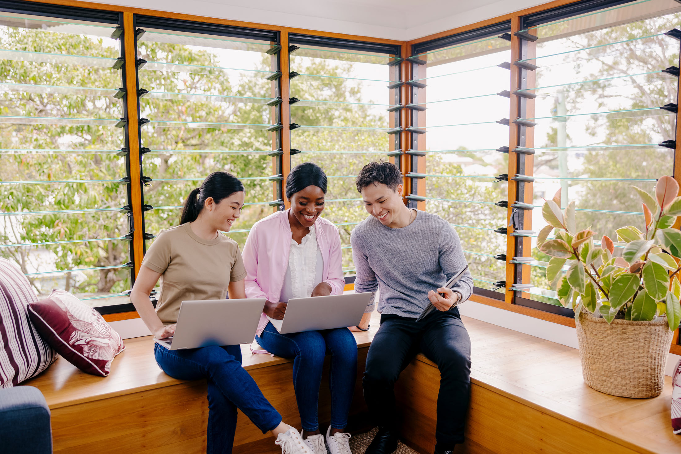 Three diverse young professionals working together on laptops in a bright sunroom with wooden bench seating and louvered windows
