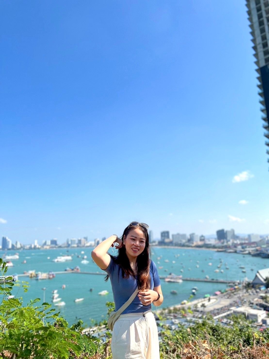 A woman stands on a hillside overlooking a bay filled with boats under a clear blue sky. The city skyline is visible in the background, and she is smiling, wearing sunglasses on her head, a blue shirt, and white pants, with a beige bag across her shoulder.