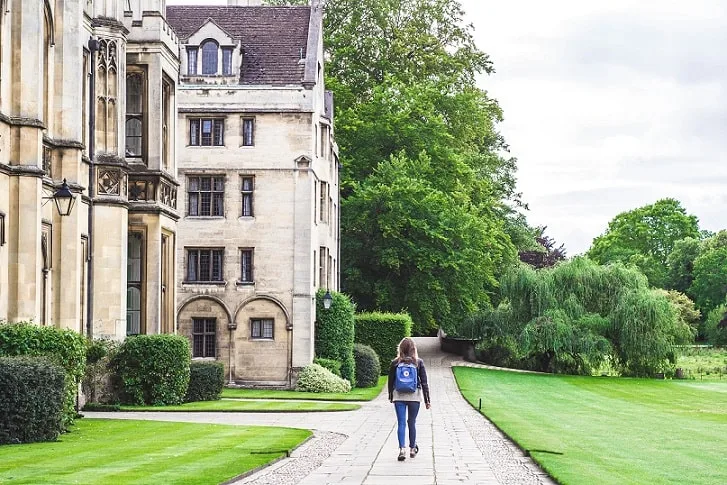 Person walking a small dog along a stone path beside manicured lawns and Gothic university buildings at Oxford College