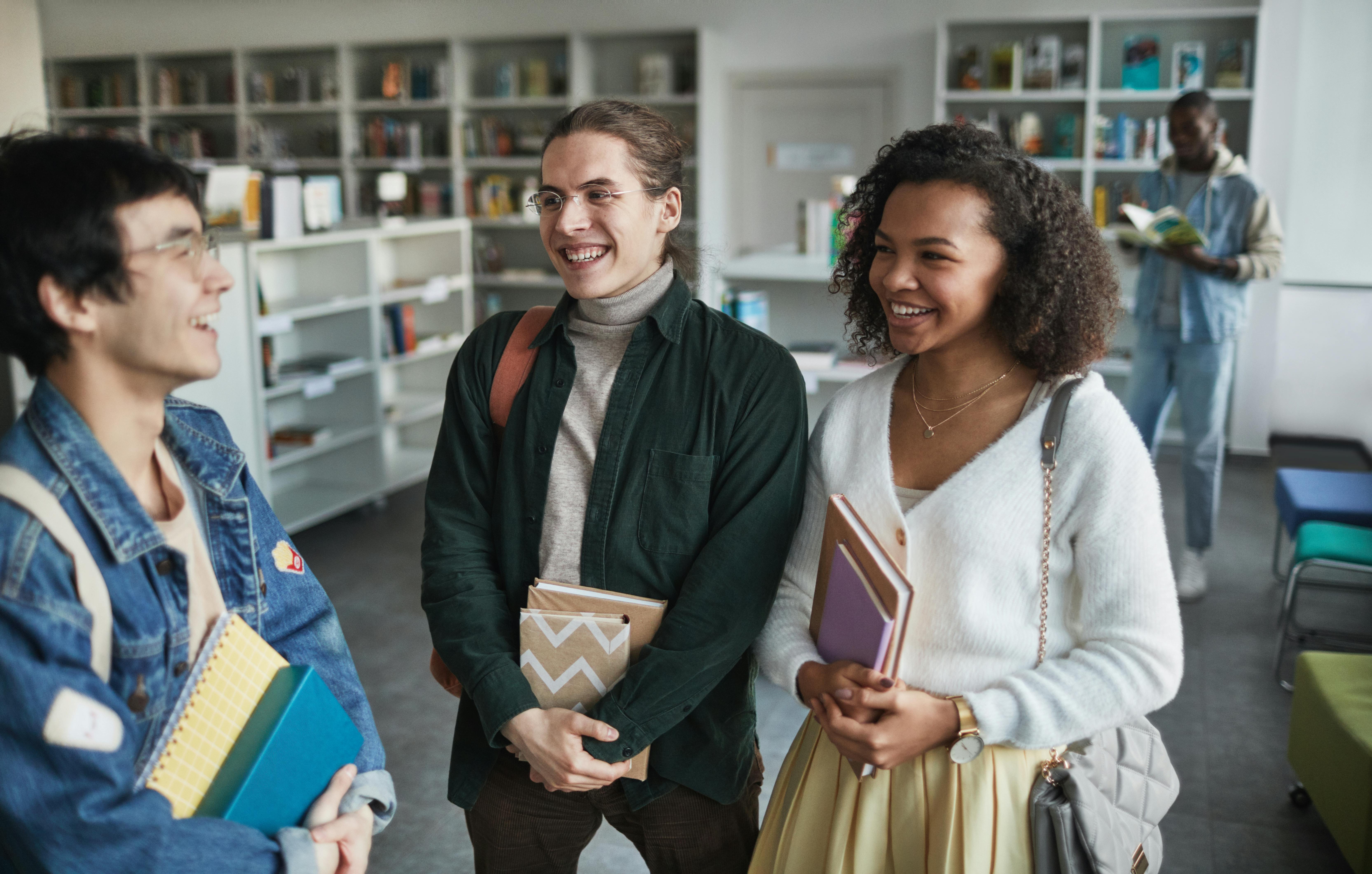 A group of three students stand in a library, smiling and holding books. Shelves filled with books are in the background, and another person is browsing. The setting is bright and welcoming.