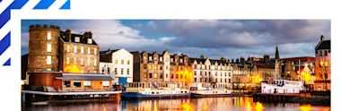 Historic waterfront in Edinburgh with boats moored along illuminated stone buildings at dusk, reflections dancing on calm waters