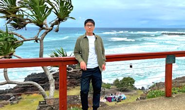 A man stands by a red wooden railing with the ocean and waves in the background. He is wearing glasses, a green jacket, white shirt, and dark pants. There is a coastal landscape with trees and people on the grass below.
