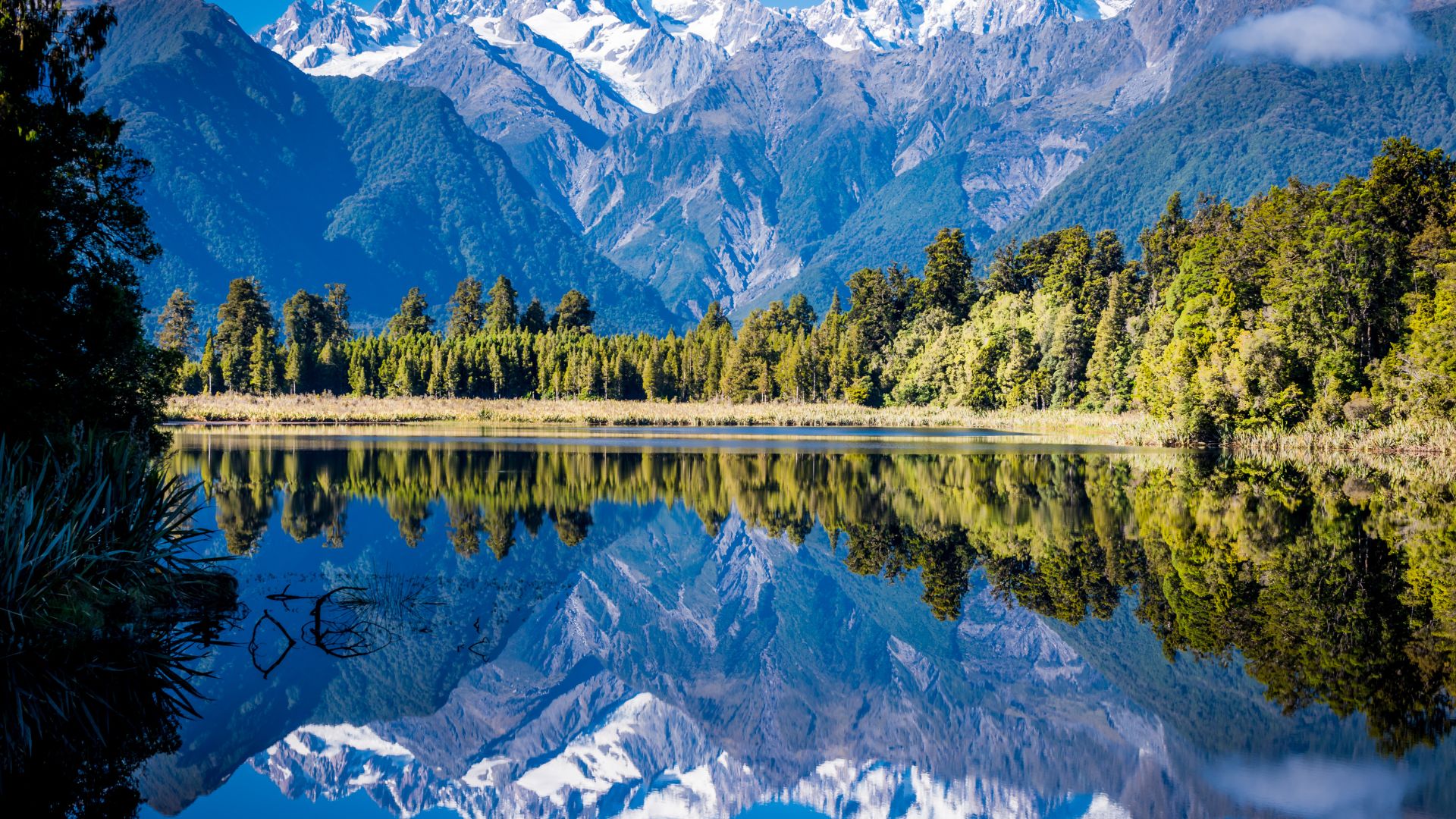 A clear lake reflects a forest and snow-capped mountains under a blue sky, creating a symmetrical, mirror-like landscape.