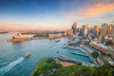 An aerial view of Sydney at sunset, featuring the iconic Sydney Opera House, the harbor, and a modern city skyline.