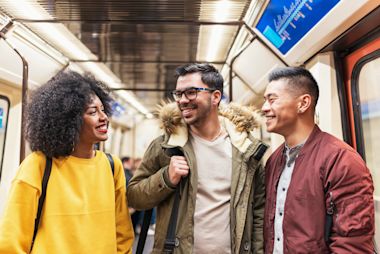 Three friends smiling and looking at each other on a train. The friend in the middle is wearing a jacket.