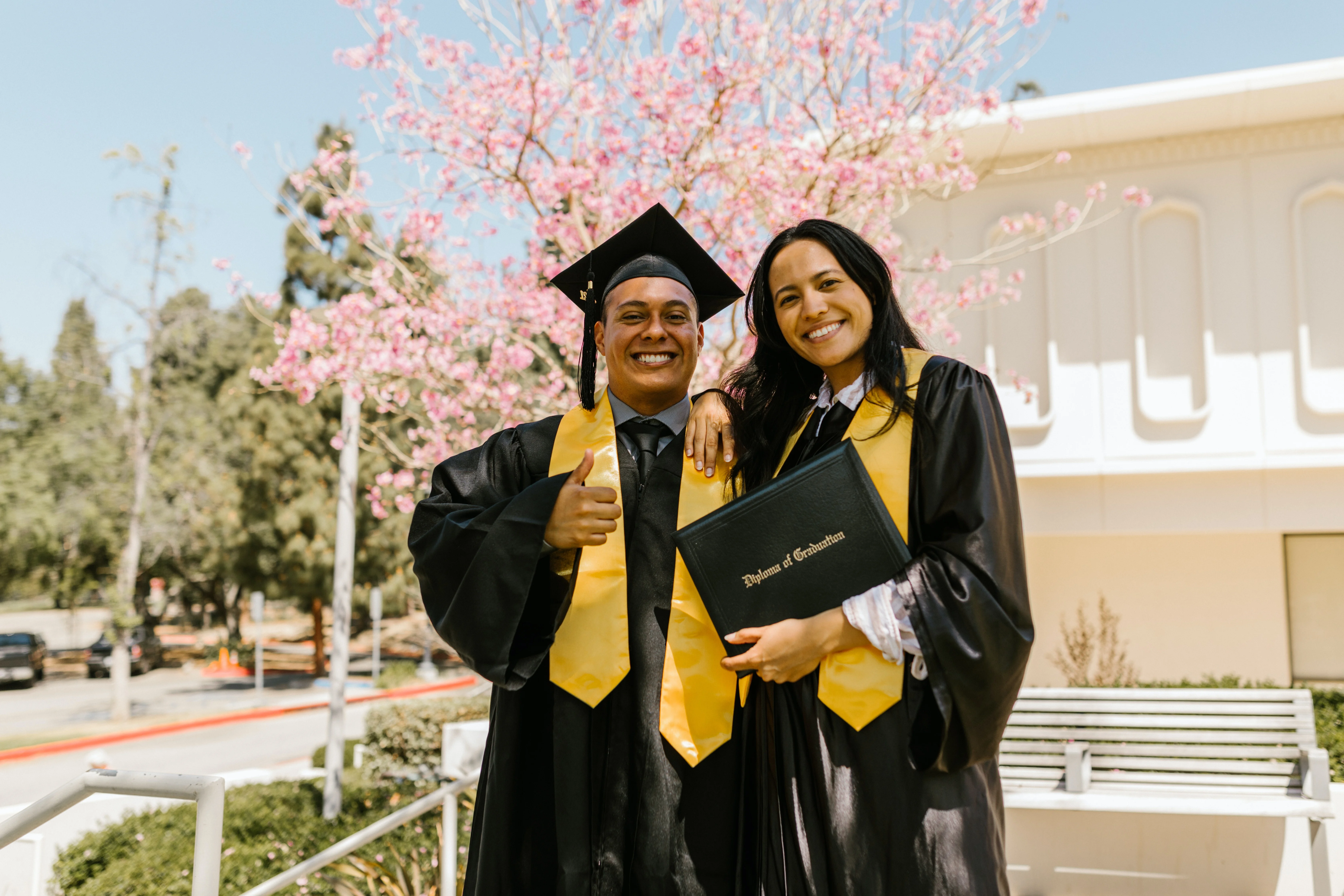 Two graduates in black caps and gowns with gold stoles pose together under blooming cherry blossoms on a sunny spring day