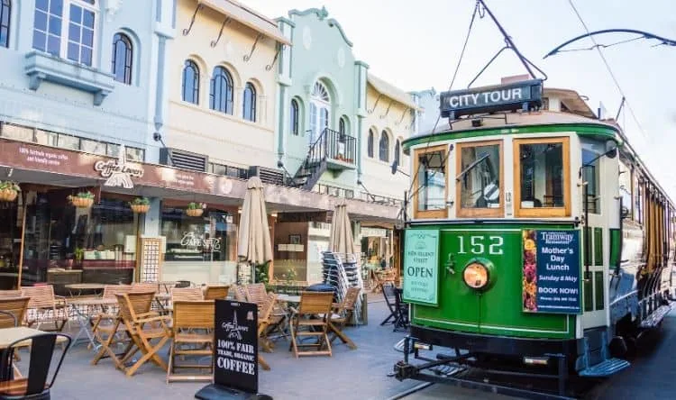 A city tour tram passing through a street where dining tables are set outdoors in front of restaurants