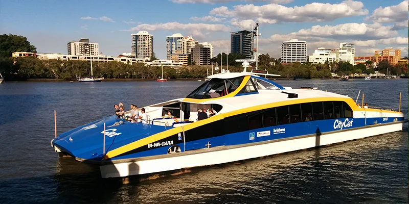 CityCat ferry cruising on Brisbane River with city skyline in background, featuring blue and yellow vessel design
