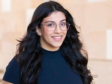 A person with long, wavy dark hair and glasses, wearing a black top, smiles while standing in front of a softly blurred, light-colored background.