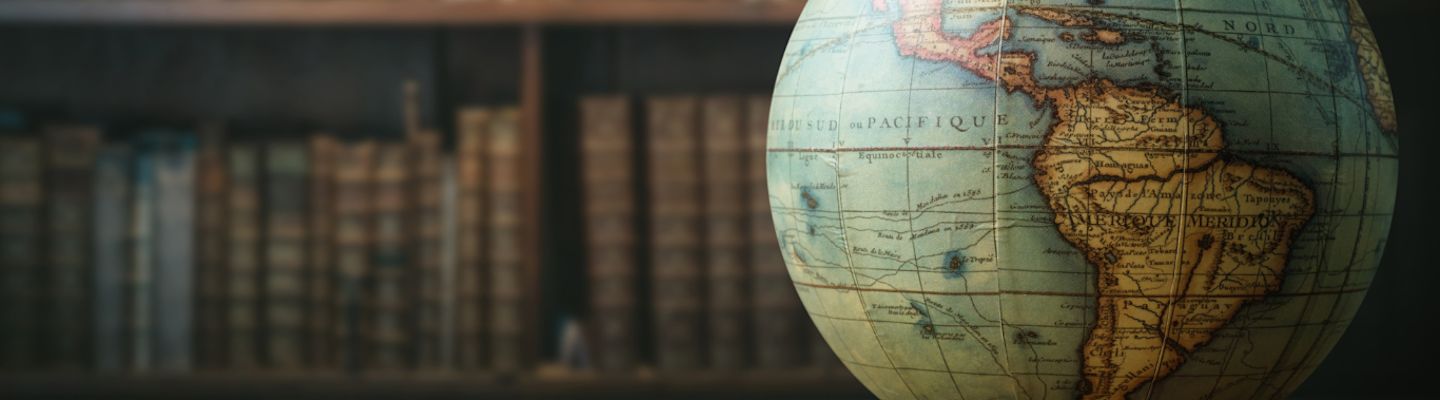 A vintage globe displaying the Americas, placed on a desk in front of a blurred background of shelves filled with old books. The globe is lit softly, highlighting its aged texture and map details.