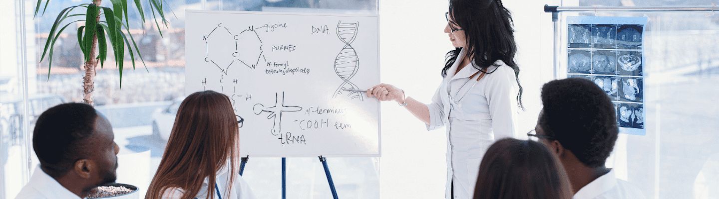 A woman in a lab coat is presenting scientific drawings on a whiteboard, including a DNA strand and chemical structures, to a group of four seated individuals in a bright room with a potted plant and X-ray images in the background.