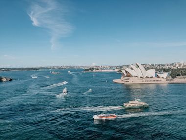  Sydney Opera House on blue harbor with ferries and boats cruising across turquoise waters under clear summer sky