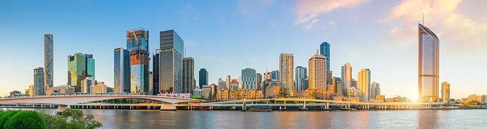 Panoramic view of Brisbane skyline with tall skyscrapers, a bridge over a river in the foreground, and a blue sky with warm sunlight illuminating the buildings.