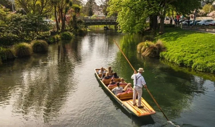 A man wearing a hat poles from the platformed till at the rear end of the boat with a group of tourists across a river