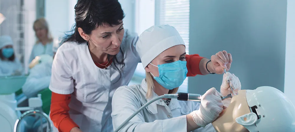 A healthcare instructor supervises a student in a lab coat and mask, practicing a dental procedure on a mannequin head in a clinical training environment.