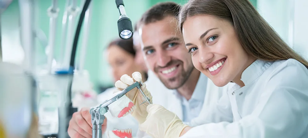Two dental professionals in white lab coats and gloves smile at the camera while working with dental models and equipment in a brightly lit laboratory.
