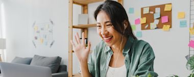 A woman waves and smiles during a video call, wearing earphones. She is sitting in a cozy room with a bookshelf and colorful sticky notes on a corkboard in the background.
