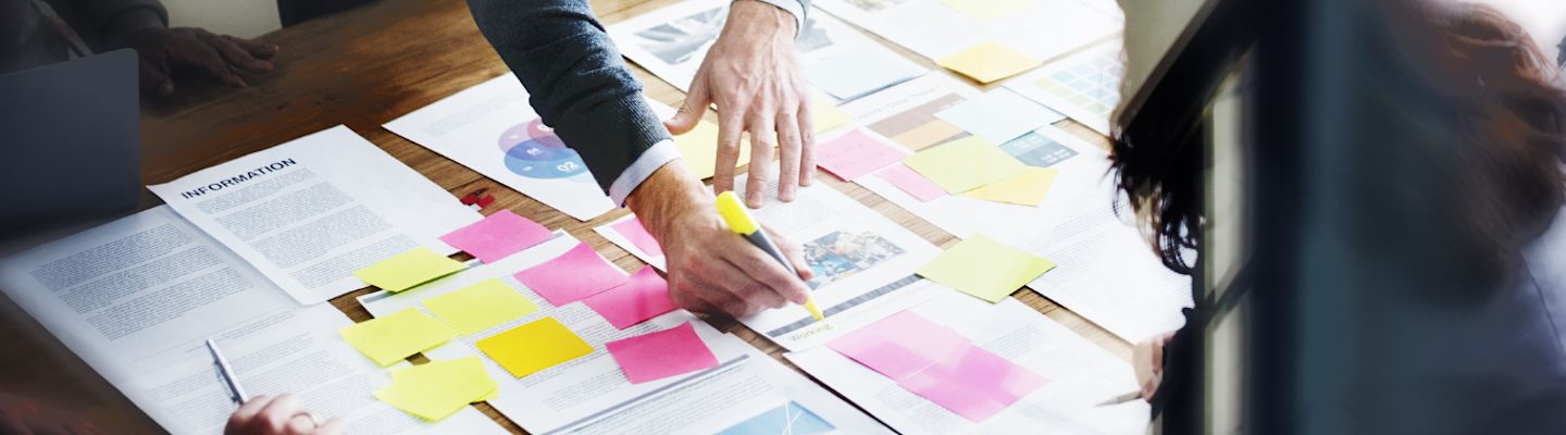 A group of people collaborating at a table filled with documents, colorful sticky notes, and a laptop. A person is highlighting text on a document with a yellow marker.