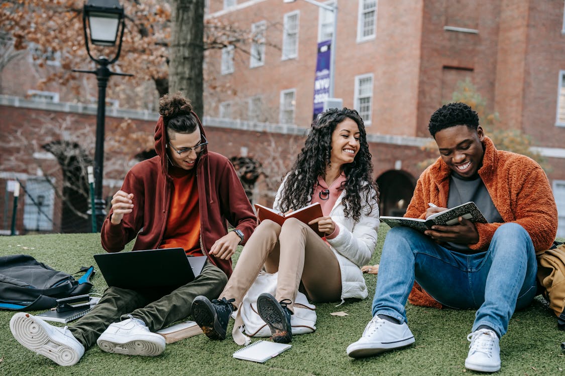 Three people sitting on grass, smiling and studying together. One uses a laptop, another reads a book, and the third holds a notebook. Theyre dressed in casual, warm clothing with brick buildings in the background.