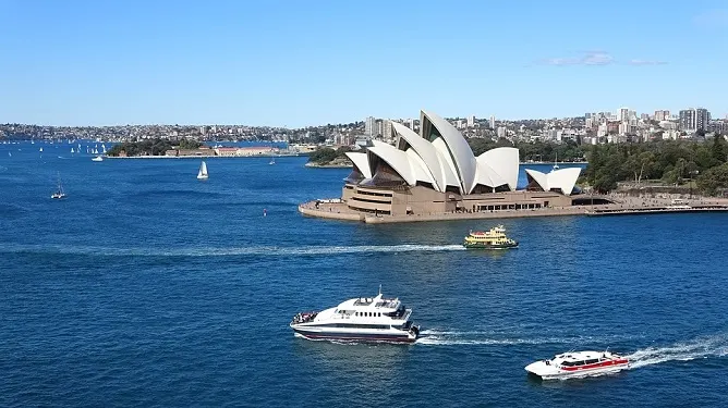 Sydney Opera House on a sunny day with ferries crossing the blue harbor waters and city skyline in background