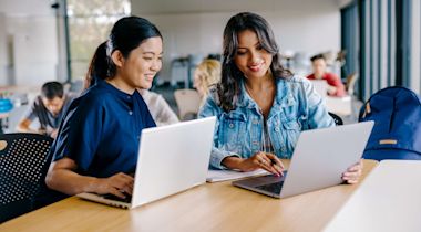 Two women sit at a table with laptops, smiling and working together in a bright, modern classroom or study area. Other students are visible in the background.