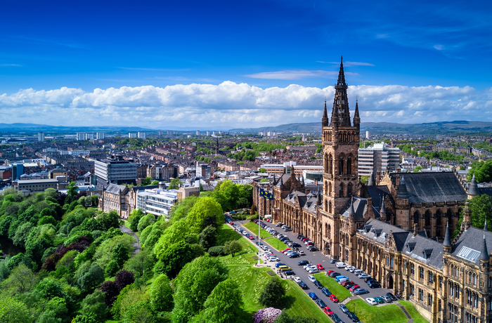 Aerial view of the University of Glasgow’s historic main building with its tall spire, surrounded by green trees, parked cars, and the cityscape of Glasgow under a blue sky with scattered clouds.