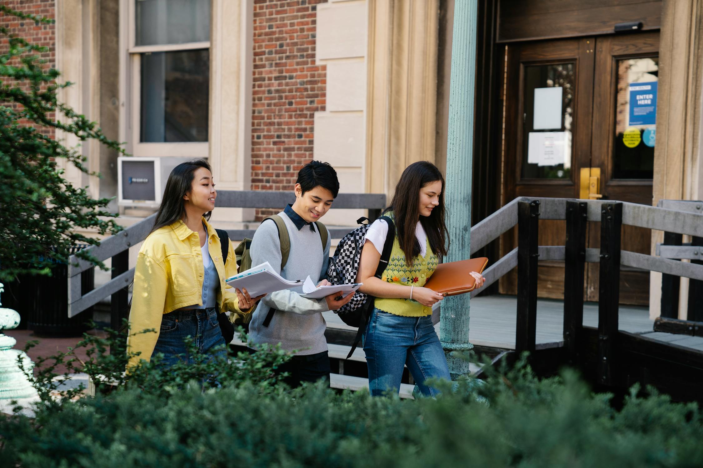 Three students walking outdoors. They are holding papers and smiling as they walk.