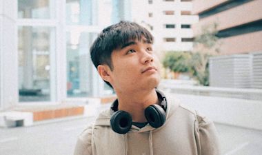 A young person with short dark hair, wearing a hoodie and headphones around their neck, looks upward thoughtfully while standing in an urban outdoor setting.