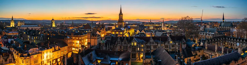 Panoramic view of a historic city at sunset, with illuminated buildings, church spires, and rooftops under a colourful sky transitioning from orange to blue. Distant hills and soft evening light frame the cityscape.