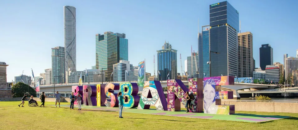 Colorful Brisbane sign with people taking photos in front of city skyline featuring modern skyscrapers on sunny day
