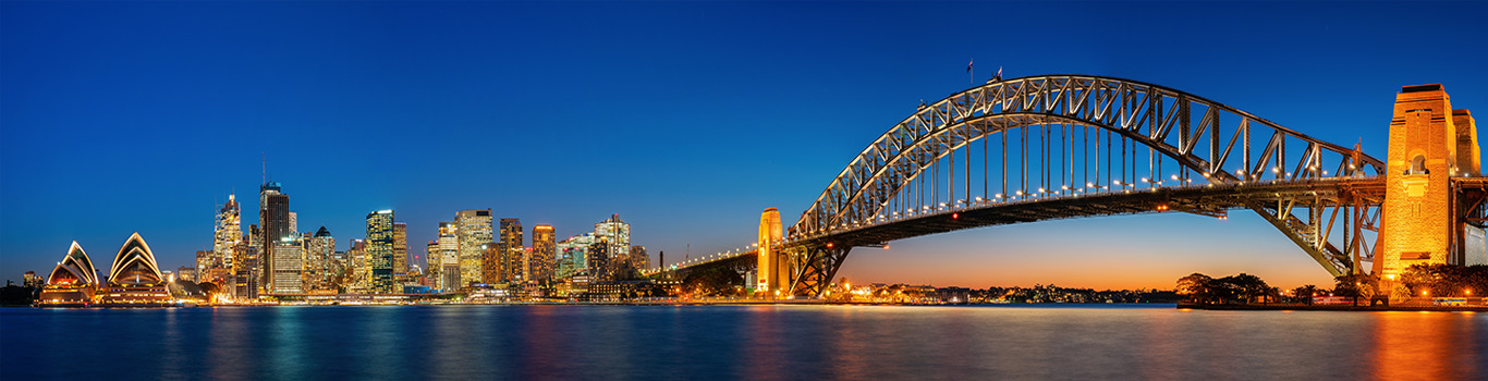Sydney skyline at dusk featuring the illuminated Sydney Opera House, city skyscrapers, and the Sydney Harbour Bridge stretching across the water, all reflected in the calm harbor under a deep blue sky.