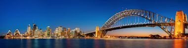 Sydney skyline at dusk featuring the illuminated Sydney Opera House, city skyscrapers, and the Sydney Harbour Bridge stretching across the water, all reflected in the calm harbor under a deep blue sky.